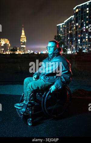 Thoughtful young man with disability in wheelchair on night walk outdoors in blue rgb neon light. Stock Photo