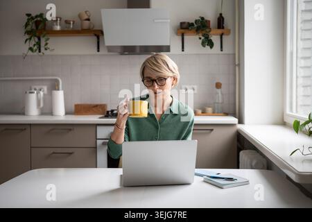 Positive middle aged businesswoman working on laptop from home and drinking coffee in early morning. Stock Photo