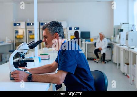 A scientist examines samples using a microscope in a well-equipped laboratory. Another researcher is seated in the background, engaged in a separate t Stock Photo