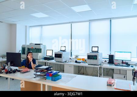 A scientist sits at a desk in a bright laboratory filled with high-tech equipment. She appears thoughtful as she reviews data on a computer screen. Su Stock Photo