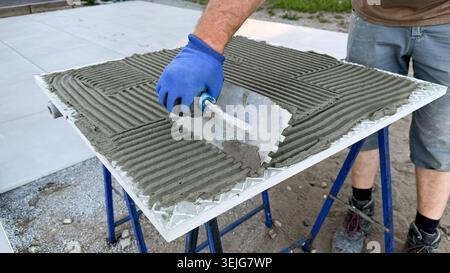 Construction worker applying adhesive mortar with a trowel on a large tile surface while standing on a support frame outdoors in a residential area Stock Photo