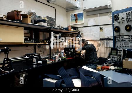 1970s England - Man working with a cine camera in an electronics laboratory, circa 1974-1976, with oscilloscopes, a reel‑to‑reel data recorder, signal generators and engineering test equipment on workbenches, UK research lab interior. Stock Photo