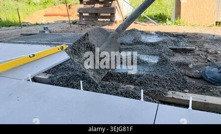 Construction worker using a shovel to pour concrete mix onto a foundation slab at a building site with wooden pallets and grass in the background Stock Photo