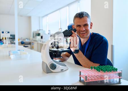 A scientist in blue scrubs is looking through a microscope in a modern laboratory. Lab equipment and samples are neatly arranged on the table, showcas Stock Photo
