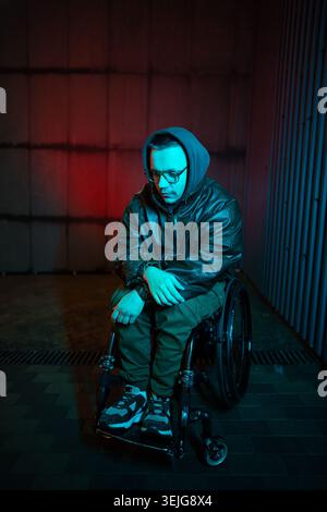 Sad man with disability sitting in wheelchair outdoors late at night under red blue rgb neon light. Stock Photo