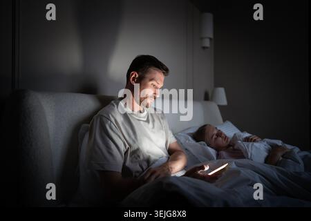 Stressed man awake at night using smartphone during sleepless hours in dark bedroom with wife. Late night phone dependency, difficulty falling asleep, Stock Photo