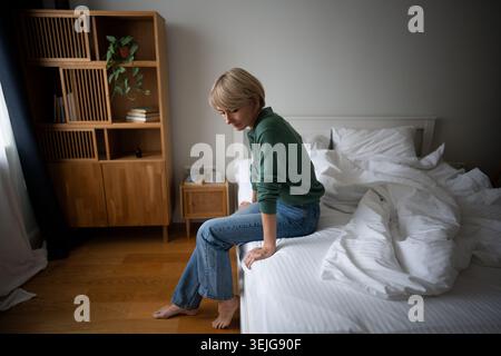 Sad tired middle aged woman sitting on bed in morning staring at side, signs of apathy and burn out. Stock Photo
