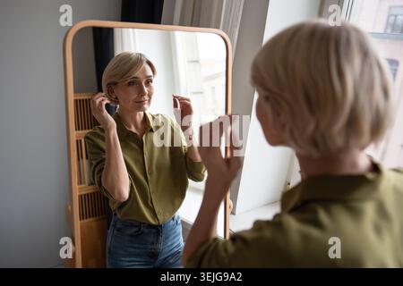 Beautiful middle aged woman looks in mirror with satisfied expression on new haircut and clothing Stock Photo
