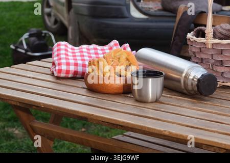 high angle view of wooden picnic table with thermos, basket and pastry near car over green grass lawn Stock Photo
