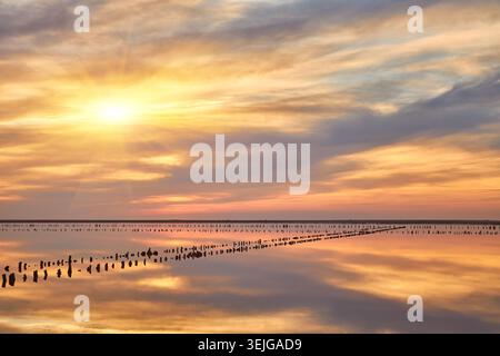 A stunning sunset over a calm salt lake. The colorful sky with orange, pink, and purple hues is perfectly reflected in the still water. Wooden posts c Stock Photo
