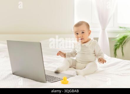 Happy baby playing, child sitting with laptop on bed in home bedroom, future IT specialist Stock Photo