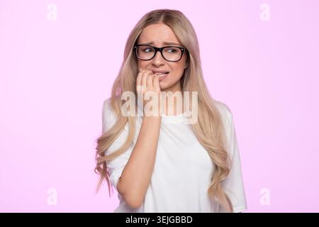 Worried young woman looks nervous, bites fingernail, feels frustrated due to an awkward situation Stock Photo