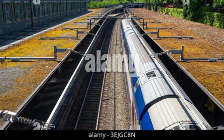 Overhead view of a blue and silver passenger train moving along railway tracks through a city station on a sunny day, with empty platforms and strong Stock Photo