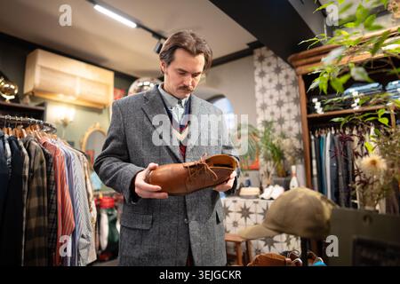 Serious handsome man customer thinking on buying fancy leather shoes in second hand vintage shop. Stock Photo