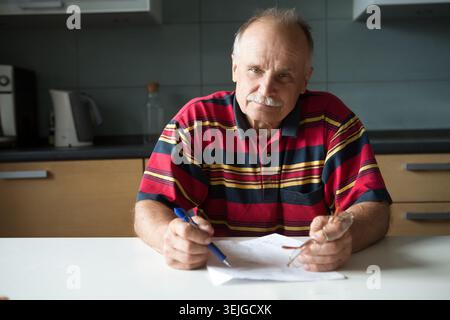 The portrait the elderly man sits at a table in kitchen.  Senior man working out he bills at home in the kitchen. Stock Photo