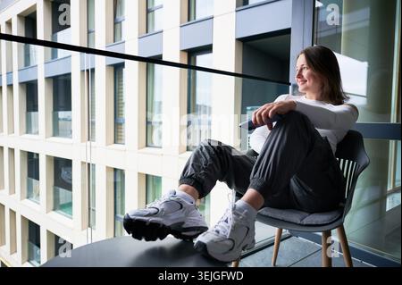 Young woman relaxing on apartment balcony with smartphone in hand. Female sitting in chair and looking away, enjoying quiet moment, urban lifestyle, comfort, residential living concept Stock Photo
