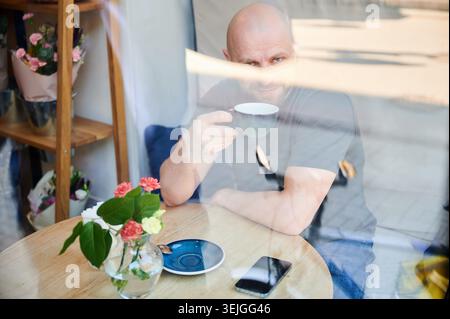 Man drinking coffee in cafe seen through window, holding cup and looking at camera, smartphone on table, reflections on glass, cozy lifestyle and urban relaxation concept Stock Photo