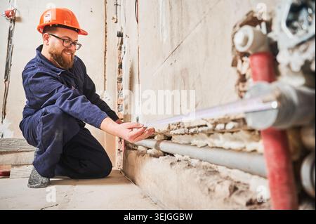 Worker measuring pipe alignment along wall near floor with tape. Smiling technician checks plumbing layout during renovation, precise installation planning and construction workflow concept Stock Photo