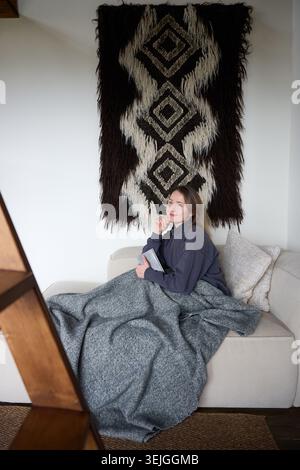 Young woman enjoying reading time on couch wrapped in blanket, cozy home setting with soft textures and minimal design, peaceful indoor relaxation concept Stock Photo