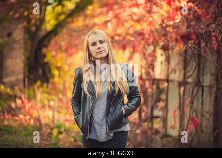Portrait of stylish young woman with blonde hair in black leather jacket. Calm and confident pose outdoors with warm autumn light and beautiful red an Stock Photo