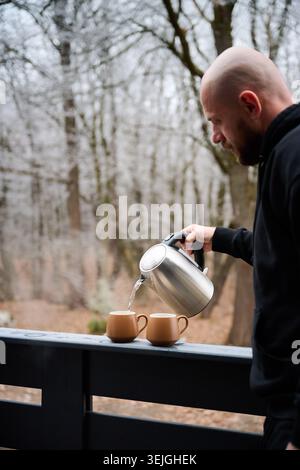 Adult male preparing warm drink with kettle on balcony surrounded by trees, symbolizing relaxation, slow living, weekend retreat, and peaceful countryside moments Stock Photo