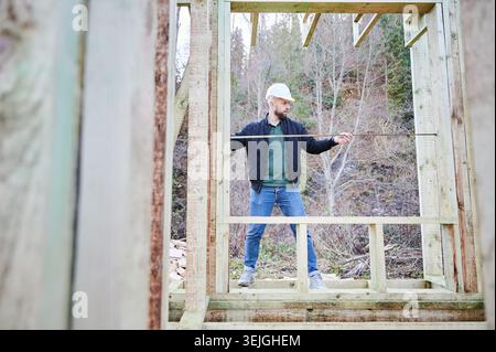 Construction worker measuring distance with tape inside wooden house frame, checking structure dimensions and alignment during building process outdoors Stock Photo