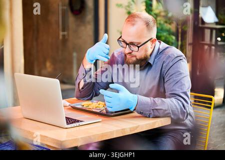 Man using laptop at outdoor cafe while eating fries, wearing protective gloves, focused on remote work and communication, concept of freelance lifestyle and modern digital routine Stock Photo