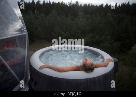 Middle aged woman rests in a hot tub in autumn cold weather against the background of green pine forest. Weekend getaway at glam Stock Photo