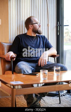 Bearded white man looking out window, seated in leather chair with remote and glasses on table, sipping water and staring thoughtfully at street view through large glass door, warm interior light Stock Photo