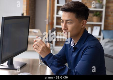 Asian man sitting at desk in office wearing navy shirt, holding glass tumbler while facing monitor. Professional, workspace, mod Stock Photo