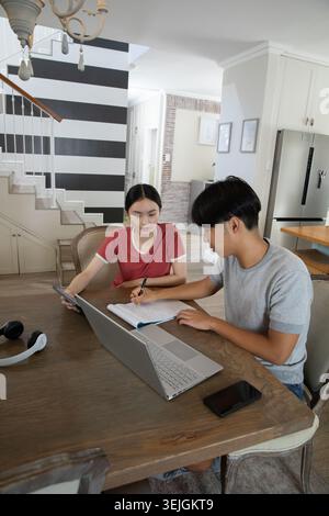 Asian students studying together at open-plan kitchen table using laptop tablet notebook headphones. Study, collaboration, learn Stock Photo