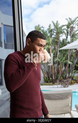 African American man in burgundy sweater leaning on door at poolside sipping coffee, copy space. Luxury, relaxation, elegance, leisure, modern, outdoo Stock Photo