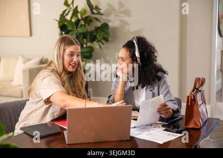 Diverse coworkers gesturing toward laptop while reviewing printed charts on table in home office. Collaboration, discussion, tea Stock Photo