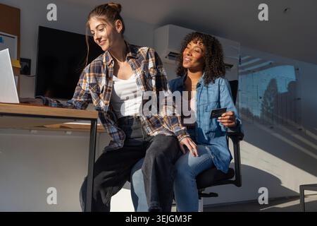 Diverse female partners typing on silver laptop while holding credit card in modern home office. Collaboration, workspace, produ Stock Photo
