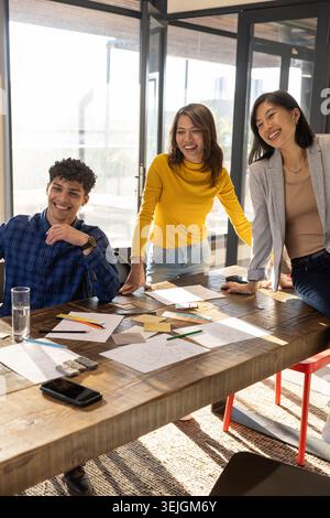 Diverse coworkers wearing business attire leaning over table in office, reviewing design sketches. Collaboration, creativity, te Stock Photo
