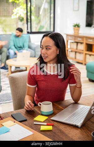 Diverse friends collaborating in living room, Asian woman in red ringer tee using laptop and mug. Collaboration, teamwork, coworking, home, bright, st Stock Photo