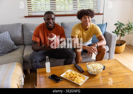 Diverse male friends holding American football while sitting on couch in living room with popcorn. Casual, friendship, relaxatio Stock Photo