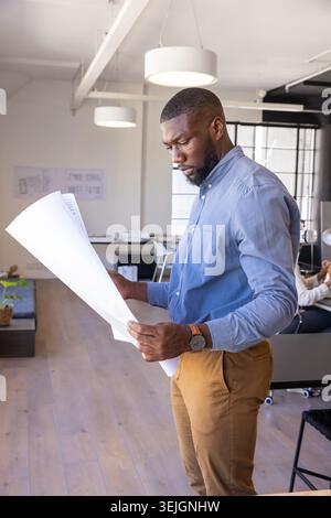 African American man standing and examining blueprints at office desk in workspace, copy space. Professional, workspace, modern, architecture, design, Stock Photo