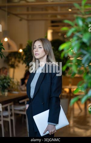 Thoughtful confident businesswoman restaurant owner posing with laptop at green terrace entrance. Stock Photo