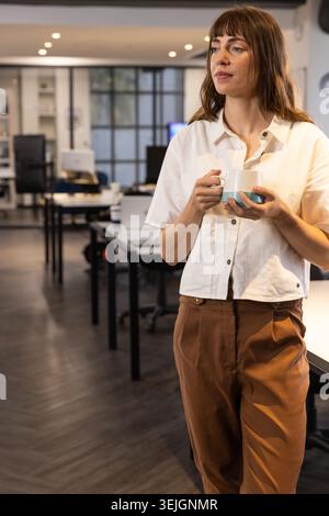 Mid adult woman wearing office outfit holding mug beside monitors in open plan office, copy space. Professional, workspace, modern, collaborative, cor Stock Photo