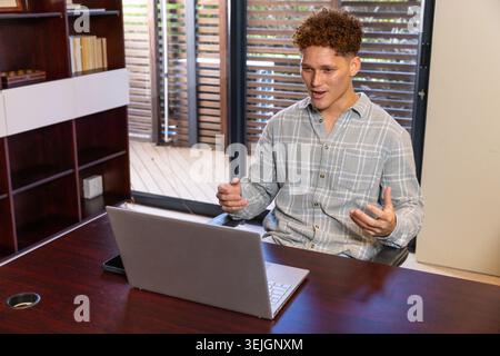 White male sitting at home office desk gesturing during online call with silver laptop. Workstation, remote work, minimalist, home environment, profes Stock Photo