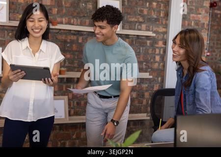 Diverse coworkers collaborating on project at desk in office with tablet, papers, pencil and laptop. Collaboration, teamwork, cr Stock Photo