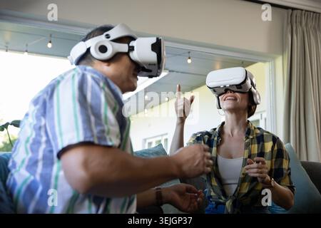 Senior couple experiencing virtual reality on two-seater sofa in living room with white VR headsets. Technology, leisure, comfor Stock Photo