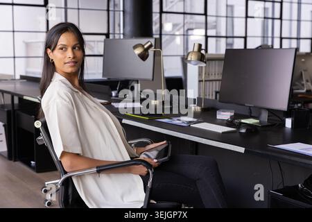 Asian woman sitting in swivel chair at open-plan office holding tablet and turning gaze right. Professional, workspace, modern, Stock Photo