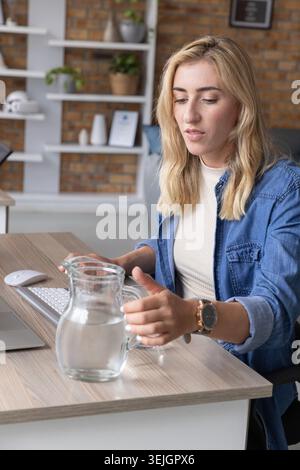 Mid-adult woman in denim shirt sitting at office desk reaching for glass pitcher near keyboard. Professional, female, blue, mode Stock Photo