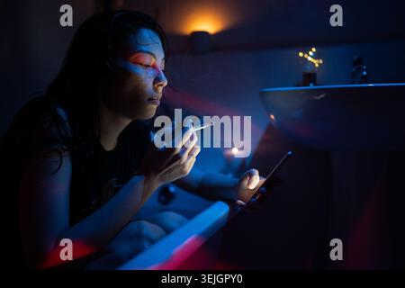 Asian woman with cigarette smoking using smartphone while sitting in bathtub under neon light, female being addicted to social networking holding cell Stock Photo