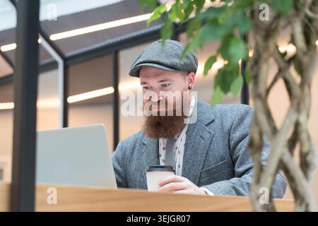 Bearded hipster man wearing wool blazer, cap working on laptop sitting in cafe Stock Photo