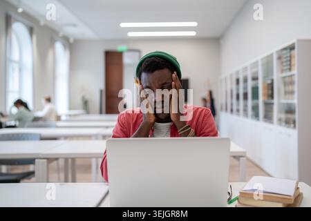 Stressed tired african american student guy tired of learning sitting in library looking at laptop Stock Photo