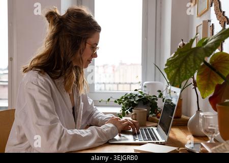 Woman freelancer/designer working on computer from home office. Cozy workplace surrounded by plants. Remote work, Telecommuting, Stock Photo