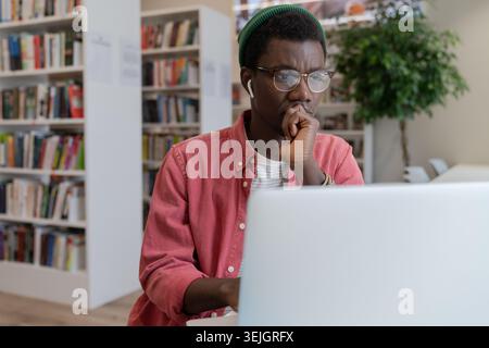 Focused pensive African American hipster freelancer man in eyeglasses working at laptop at library workplace table thinking over IT online startup pro Stock Photo